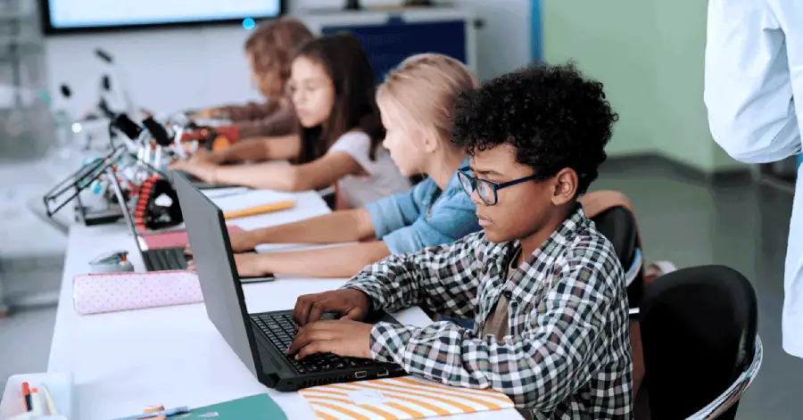 Side view of a schoolboy and his classmates working in front of laptops while sitting in a row by desks, highlighting accessibility in K12 education. 