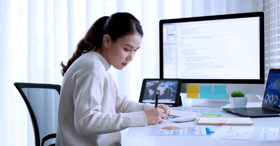  A young Asian businesswoman sits at her home office desk, utilizing both a desktop and laptop, contributing to her own workforce development. 