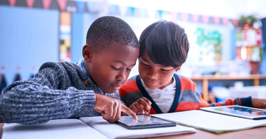  Two young boys using a tablet reading benefits of AI in education in a classroom. 