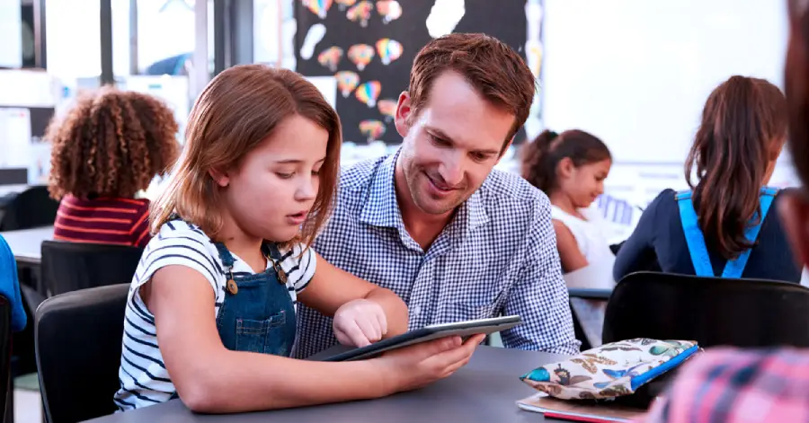  A male teacher and a young girl using a tablet in a classroom reading the benefits of AI in education with other students in the background.