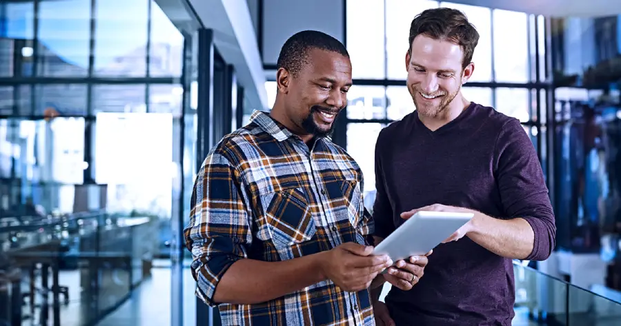 Two men standing in a modern office environment, smiling and looking at a digital tablet together reading the 10 latest Learning and Development trends.