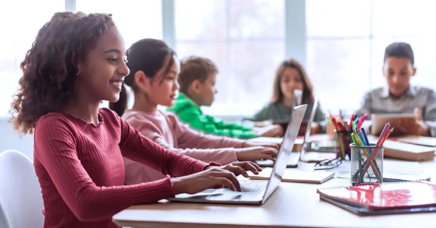 A black schoolgirl using a laptop sitting at a desk with multiethnic classmates in a classroom.