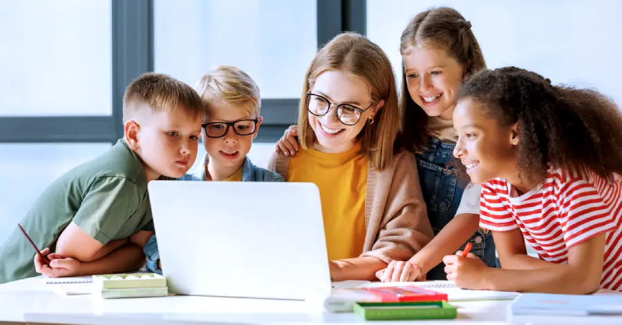 A multi-ethnic group of students learning from a laptop with a female teacher wearing specs teaching them about the benefits of custom content for K-12 schools. 