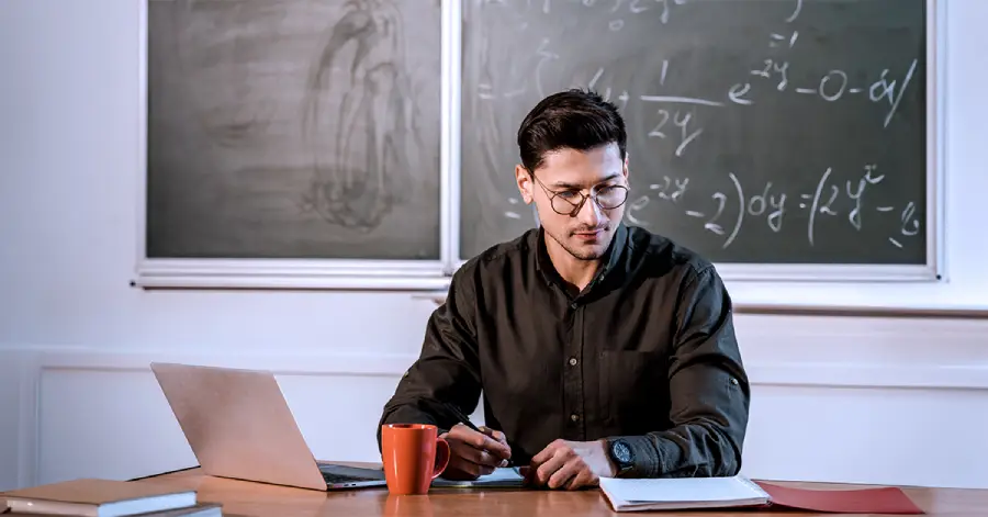  A male teacher wearing specs with his laptop and a notebook, searching apt tools for custom content development.