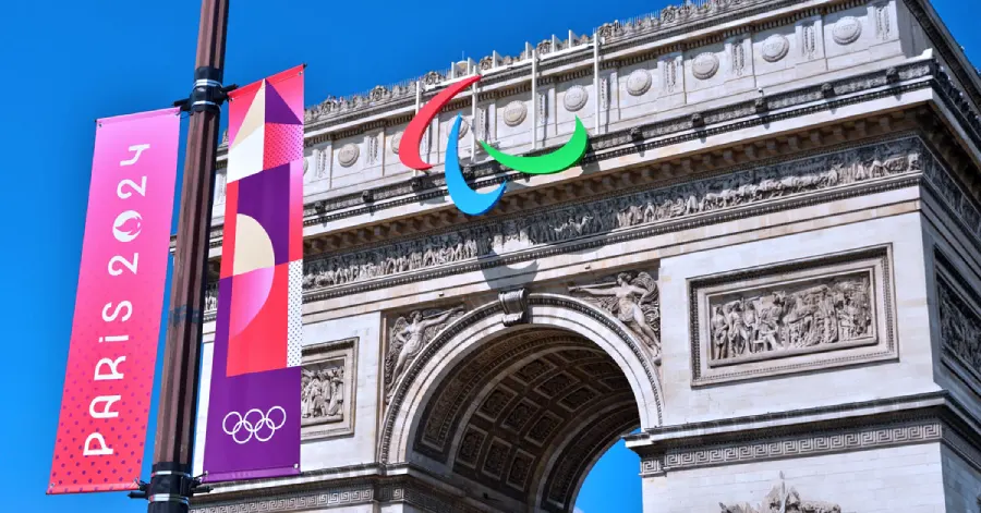 An image of The Arc de Triomphe with the Paralympics symbol on it. The flags of the Paris Olympics 2024 are on a poll in front of it. 