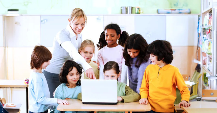 A teacher guiding a diverse group of young students gathered around a laptop in a bright, colorful classroom.  
