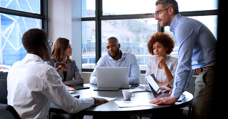 A diverse business team collaborates during a meeting in a modern office space. Five professionals sit and stand around a table with a laptop, documents, and coffee cups. The office features large windows with natural lighting.