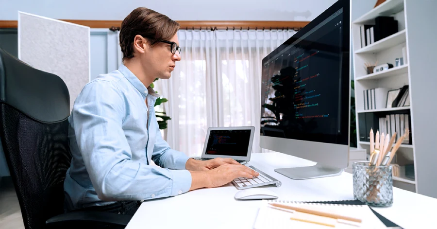  An  IT developer in a light blue shirt and glasses working at a home office setup with dual monitors, while a laptop sits beside it on a white desk.
