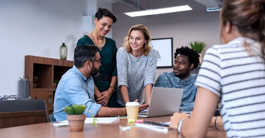 An image of multicultural working professionals in an office setting, with a laptop, coffee cups, and plants visible. 