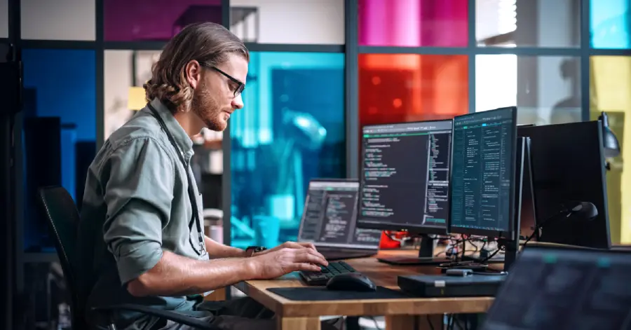 An image of a male programmer on a desktop computer with multiple monitors, working in an office environment. 